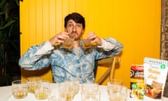 A man sitting at a table, holding two glasses, looking unsatisfied. In front of him are many glasses filled with chicken stock.