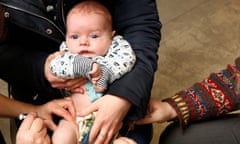 A baby receives a routine two-month vaccination against whooping cough and other conditions including diphtheria, tetanus and polio at a health centre – he is cradled in his mother's arms while a nurse injects a syringe into his thigh and the arm of the doctor is supporting