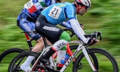 Mohammad Ganjkhanlou in a cycling race – he is slightly ahead of another rider whose head is not seen; he wears a light blue top with white long-sleeved underlayer and black shorts over green and white leggings; he rides a white bicycle