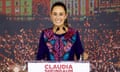 A slim, middle-aged Latina woman with pulled-back brown hair and a color long-sleeved top in purple, blue, pink, and red, stands at a lectern that has her name on it, smiling.