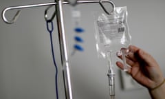 A hand reaches up toward a clear IV drip bag suspended from a metal tower.