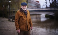 Man wearing cap and brown jacket stands next to a river with a bridge in the background