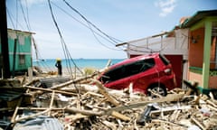 A car rests at a 45-degree angle to the ground partially obscured by surrounding broken timber, detritus and downed power lines.