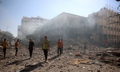 a group of Palestinian men walk on the rubble and debris of a school destroyed by an airstrike; other damaged buildings and smoke can be seen in the background under a blue sky.