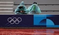 People wearing raincoats sit in Roland Garros Stadium, which has been decorated with the Olympics rings logo