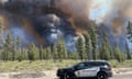 A black and white police SUV parked on a sunny road alongside trees, with endless smoke rising beyond it.