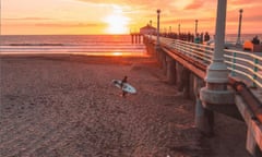 A man carries a surfboard along Manhattan Beach, California, above him, on a pier, people promenade as the sun sets.