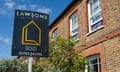 A dark green sold sign with yellow lettering and house symbol outside an old brick house with decorative brick detailing around its windows and white wooden windowframes; there is greenery and a tall bush in the front garden behind the sign; the house appears to be quite small and in a terrace but looks smart and desirable