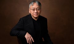 Kazuo Ishiguro, in a T-shirt and blazer and wearing glasses, smiles with his arm on the back of a chair armrest