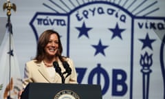 Black and South Asian woman with shoulder-length brown hair smiles and speaks at lectern in front of white and blue logo behind her.