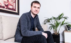 Michael Robinson, with short hair and wearing a zip cardigan, T-shirt and jeans, sits on a sofa next to a palm plant and smiles slightly