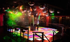 A dark nightclub with an illuminated multicolour dance floor and a man standing next to a mirrored pillar