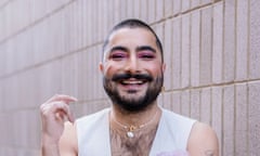 Person with a beard, moustache and pink eyeshadow, dressed in an off-white waistcoat, standing against a concrete-tiled wall