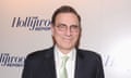 Older white man, dyed brown hair combover, glasses in black suit with pale green tie in front of press background that says "The Hollywood Reporter."