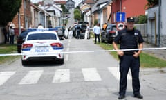 A police officer stands behind police tape cordoning off a road. Behind him on a residential street is a police car and people putting on forensics suits