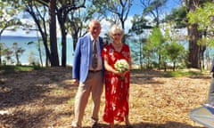 A couple aged in their 70s on their wedding day. The man is wearing a light-blue jacket and beige trousers, the woman is wearing a red floral dress and holding a bouquet of flowers. They are both smiling