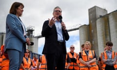 Keir Starmer speaks into a microphone. Rachel Reeves stands beside him. Behind them are people wearing orange hi-vis vests