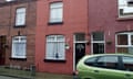 A child pulls aside a net curtain as they look out the window of the red-brick terrace house, which has one upstairs and one downstairs window at the front and a black front door