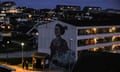 A mural of a woman and a polar bear on the side of a block of flats in an urban area lit up after dusk