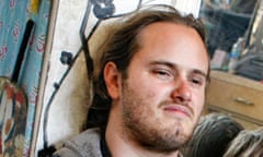 A 20-something white man with scraggly beard and long dark blond hair, appearing to sit on a floor leaning against a doorway, smiling slightly.