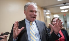 Older white man in hallway among reporters, hands raised in gesture, speaking, wearing a suit and yellow tie.