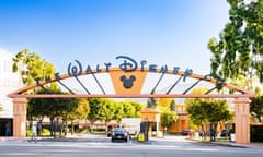 the entrance to Walt Disney Studios and corporate headquarters in Burbank, California: a wide driveway is seen under an arch with lettering reading The Walt Disney Co and a symbol in the shape of Mickey Mouse's head and ears. A car is coming under the arch, which has trees to either side and in an avenue leading to buildings; it looks hot and sunny with a clear blue sky above.