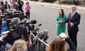 Swinney in suit and Forbes in green dress talking to dozens of journalists with mics and cameras behind a barrier