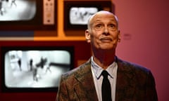 An older white man, mostly bald but also with very close-cropped white hair and a drawn-on black line as a moustache above his upper lip, look up toward a light that is shining on him. He stands before a pink wall that has a bank of black-and-white TVs, and wears a paisley suit coat, a white collared shirt, and a skinny black tie.