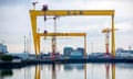 two huge yellow cranes loom over the water at the shipyard with industrial buildings in the background against a grey sky