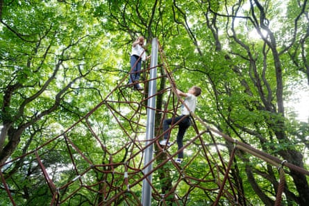 Two children climb a rope climbing frame beneath large verdant trees.