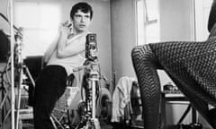 David Bailey, in a vest and tight trousers, hands clasped, bending a little backwards, with Jean Shrimpton's leg resting on a table