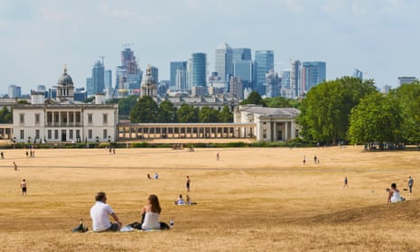 People sit on parched grass in Greenwich Park in summer looking north towards Canary Wharf