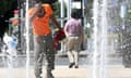 A man in an orange shirt takes off his construction helmet in order to splash water on his head in a public fountain