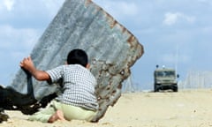 A Palestinian child kneels behind a sheet of corrugated iron as they look at an armoured vehicle