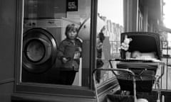 A black-and-white photo of a child standing beside a washing machine in a laundrette, with a baby in a pram just outside the window