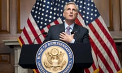 Kevin McCarthy with his hand on his heart standing at a podium with a House of Representatives seal on the front of it and the US flag in the background