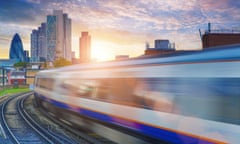 A blurred, speeding London overground train hurtles towards the skyscrapers of the City of London, including the Gherkin, as the sun rises in the background