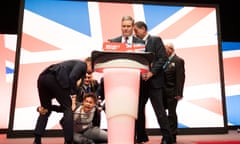 A shouting protester kneels on the floor next to a glitter-covered Keir Starmer. The protester is holding the leg of one of five people trying to get him off stage.
