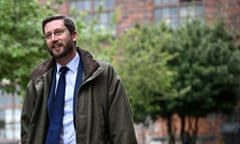 Simon Case, seen outdoors and wearing a waxed cotton jacket, pale blue shirt and dark blue tie; he has a thick but neat beard; there are brick buildings and trees seen in the background and he is smiling.