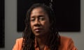 A Black woman with long thin dreads and an orange blazer looks seriously at the camera in a portrait.