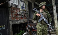 An armed soldier in military fatigues stands next to a wall of graffiti.