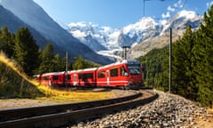 A red passenger train passing pine trees, with snow-capped mountain in the background