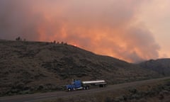 A large lorry drives by a hill with smoke from a wildfire rising from the other side
