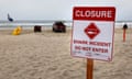 Red and white sign stapled to wooden post put in sand says Closure, Shark Incident, Do Not Enter, with date handwritten in ink, beyond which is red truck facing ocean and one person walking.