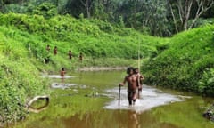 Seven men from an Austroasiatic Indigenous group wearing loin cloths ad carrying sticks or spears walk along a river in a forest