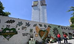 People next to a Grenfell tower fire memorial wall, with the tower's tarpaulin-wrapped ruins visible in the background