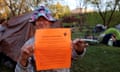 Weathered-looking white woman wearing pink and blue sunhat holds up bright orange piece of paper, with tents on green grass behind her.