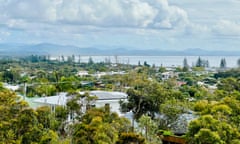 View of Byron Bay, with trees, rooftops, trees and the distant ocean and hills under a cloudy sky