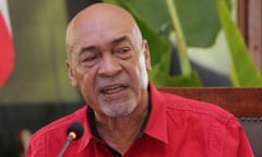 A bald man wearing a collared red shirt speaks into a microphoned as he is seated at a table with large green plants in the background.