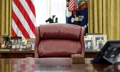 A big cushy brown leather chair sits behind a wide desk in what is assumed to be the Oval Office, with a window with long yellow curtains and an American flag behind it, beyond lots of framed photographs on a bureau top.